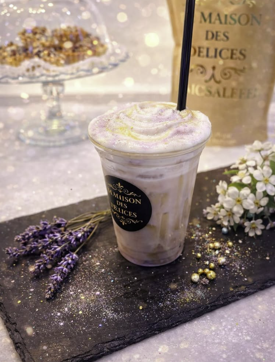 Ein geeister Ube Latte mit dem ‚La Maison des Délices‘-Logo auf einer schwarzen Schieferplatte, dekoriert mit getrocknetem Lavendel und weißen Blüten sowie einer lila-gold glitzernden Creme-Haube.