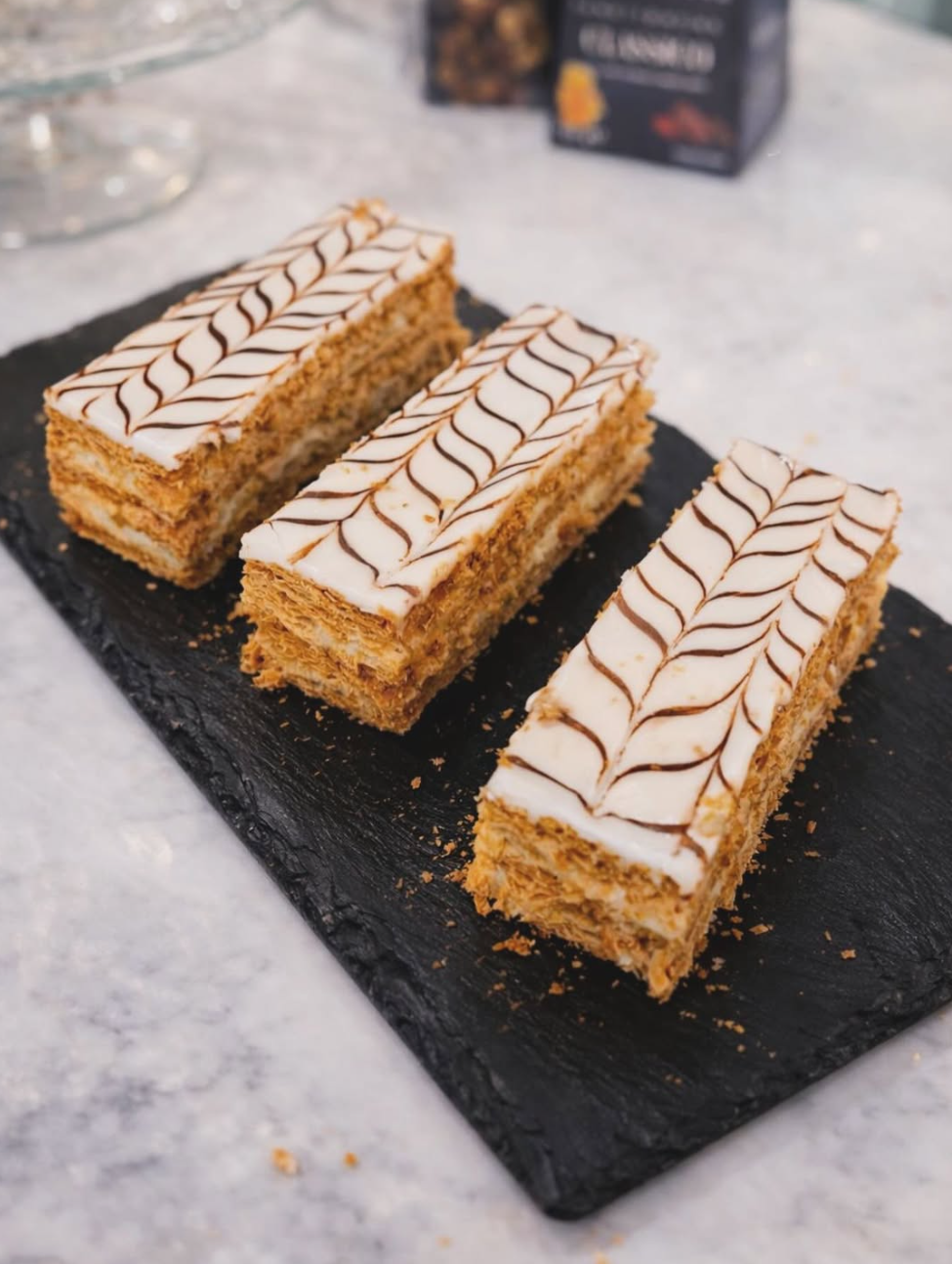 Drei frisch zubereitete Mille-feuilles auf einer schwarzen Schieferplatte. Zu sehen sind goldbraune Blätterteigschichten, eine weiße Cremefüllung und die charakteristische braune Zickzack-Verzierung auf der glatten Zuckerglasur.