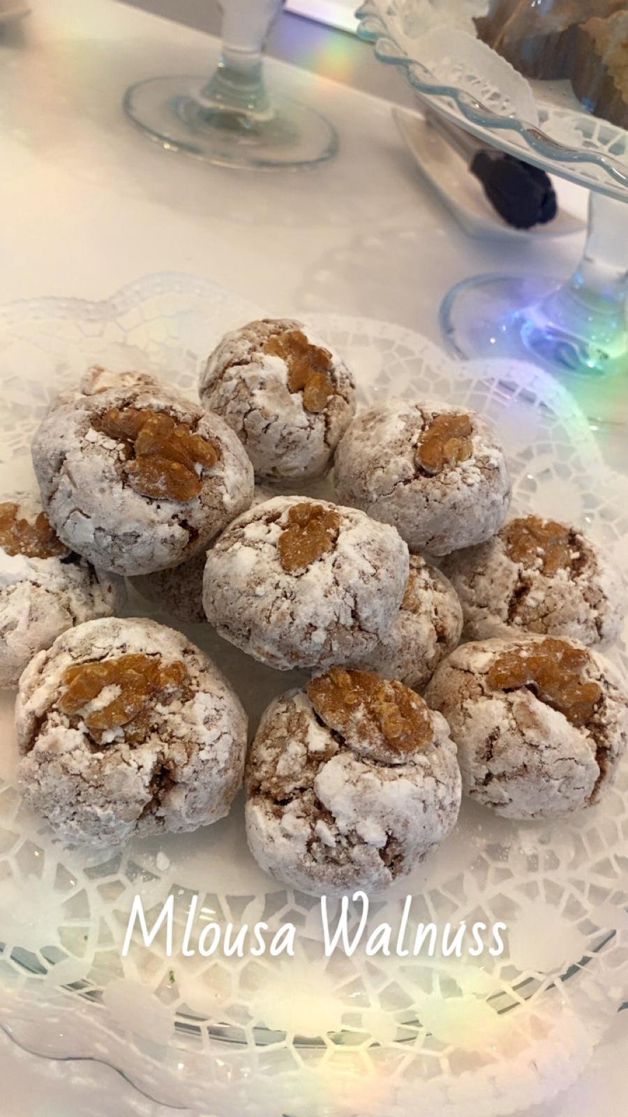 Auf einem Papier mit weißem Spitzenmuster präsentierte "Mlouza Walnuss"-Kekse, die mit Puderzucker bestäubt sind und in deren Mitte jeweils eine ganze Walnuss platziert wurde.