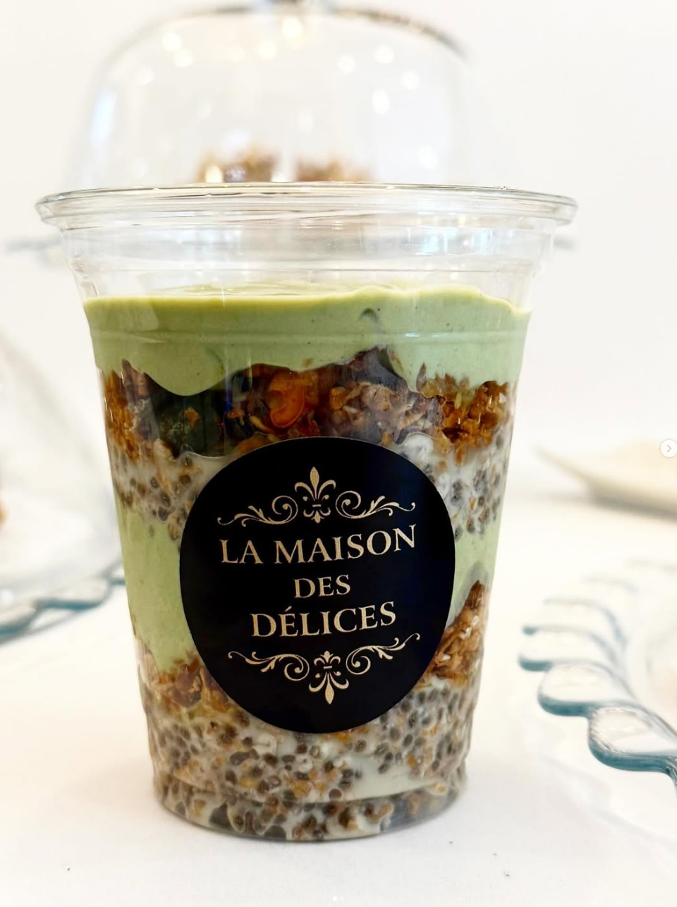 Ein gesunder Snack in einem transparenten Behälter, geschichtet mit Chia-Pudding, Granola und Matcha-Creme, versehen mit dem ‚La Maison des Délices‘-Logo.