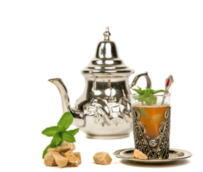 Vor einem weißen Hintergrund steht eine traditionell verzierte silberne Teekanne (Berrad), daneben ein Glas goldfarbener marokkanischer Tee, der mit frischen Minzblättern garniert ist.