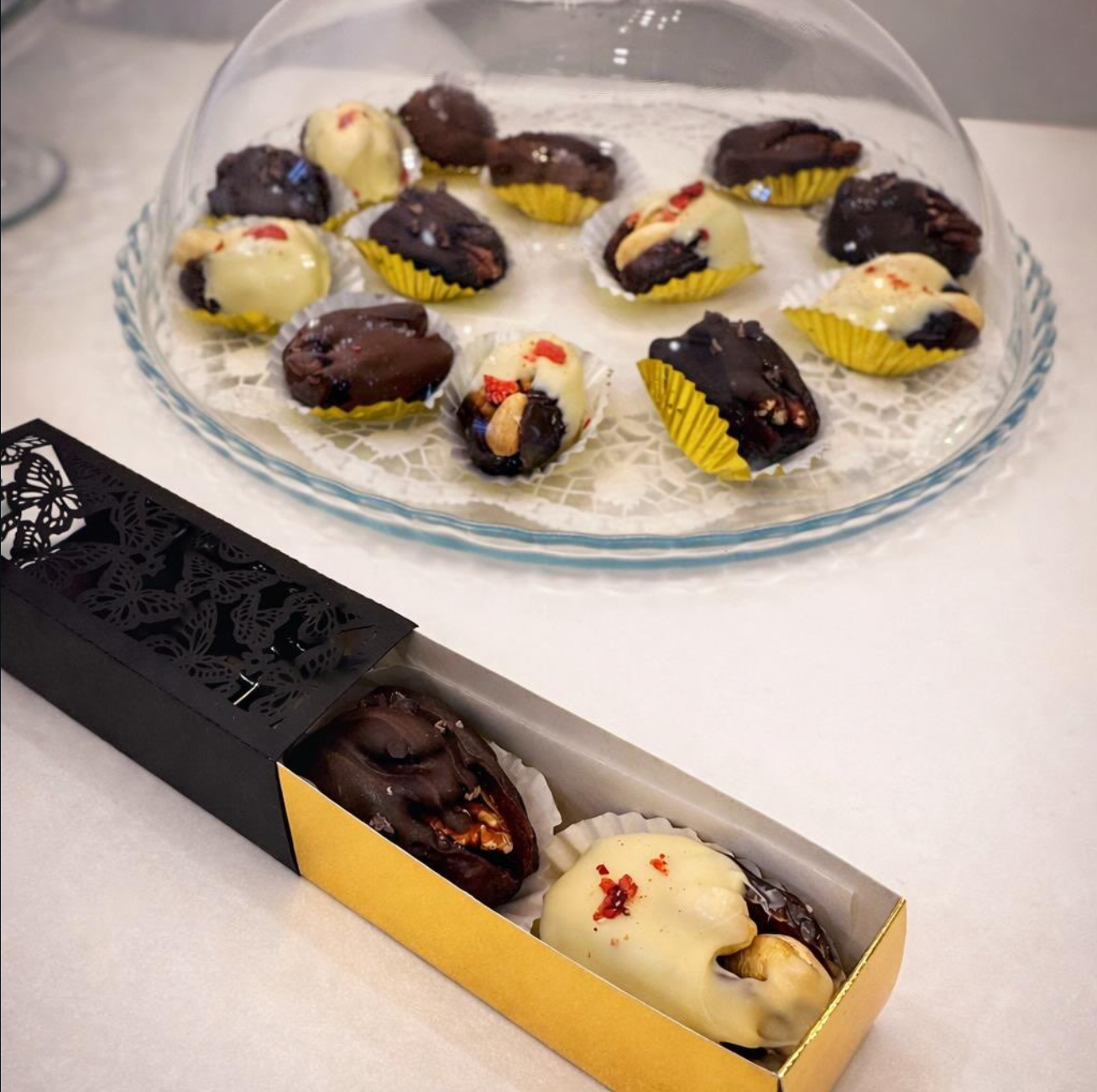 Beschreibung für Barrierefreiheit und SEO: Eine elegante schwarz gemusterte Box mit goldenen Akzenten, in der zwei gefüllte Datteln nebeneinander liegen; im Hintergrund eine edle Glasplatte mit Medjoul-Datteln, die mit weißer, Vollmilch- und Zartbitterschokolade überzogen und mit Cashews, Pekannüssen sowie Erdbeerstücken veredelt sind.